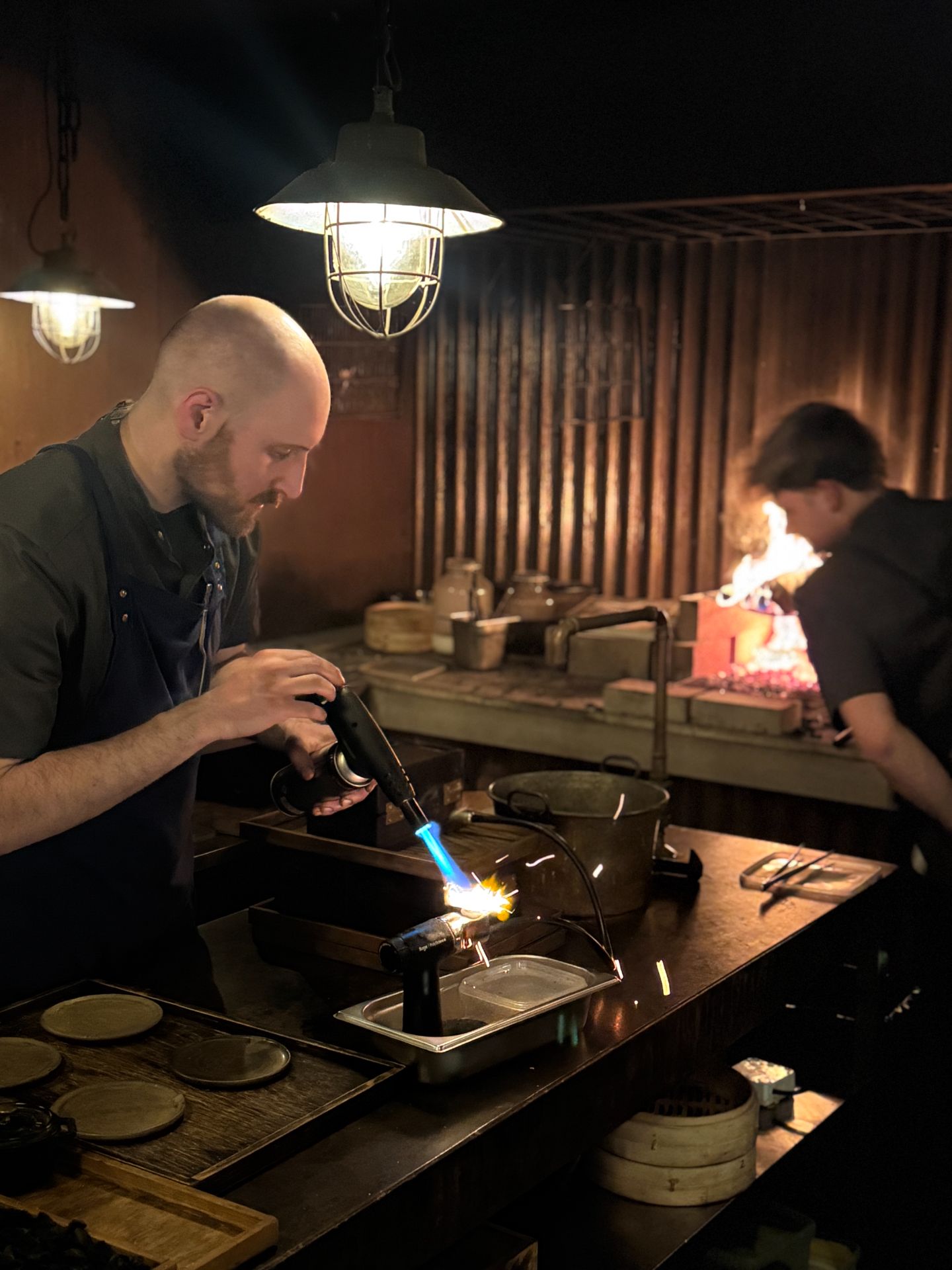Two chefs at a kitchen counter, one using a blowtorch, with a wood fire burning behind them.