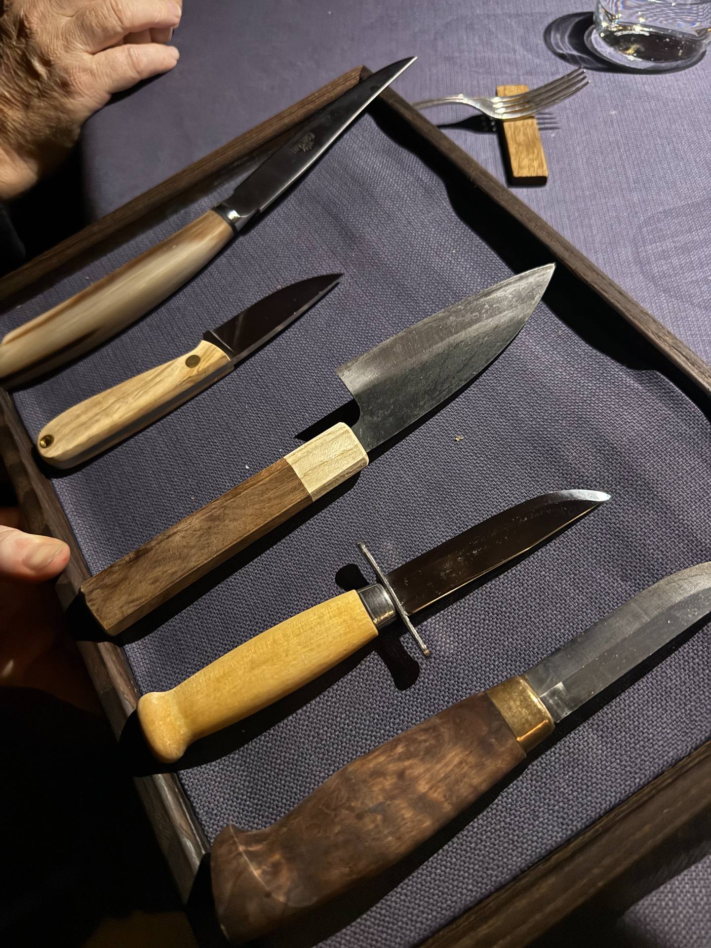 Five knives with different handles displayed in a wooden frame on a purple tablecloth.
