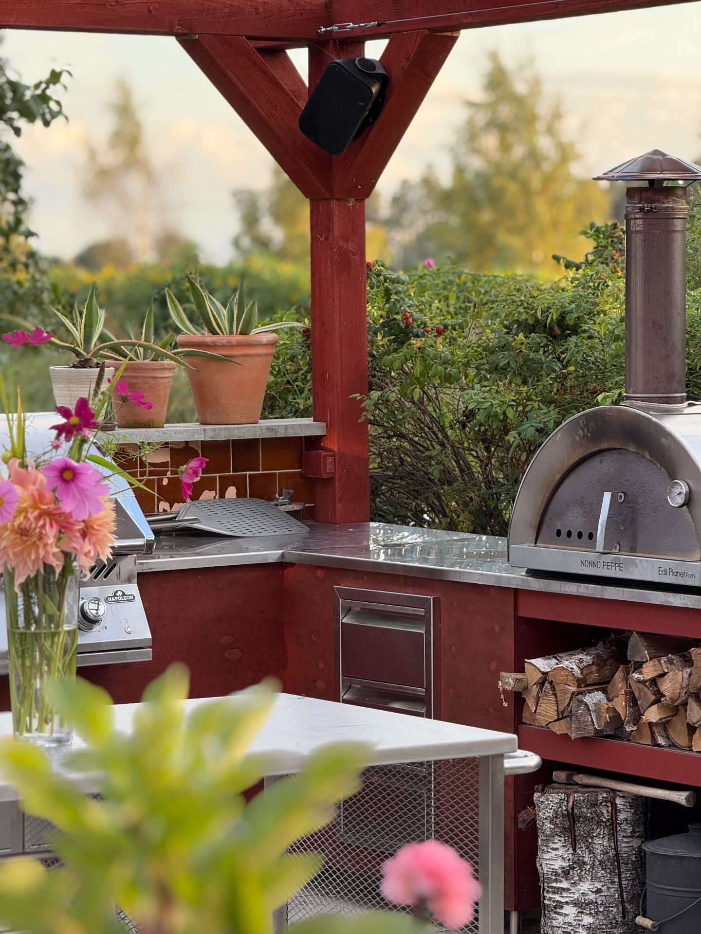 Outdoor kitchen with a pizza oven, stacked logs and flowers beneath red beams.