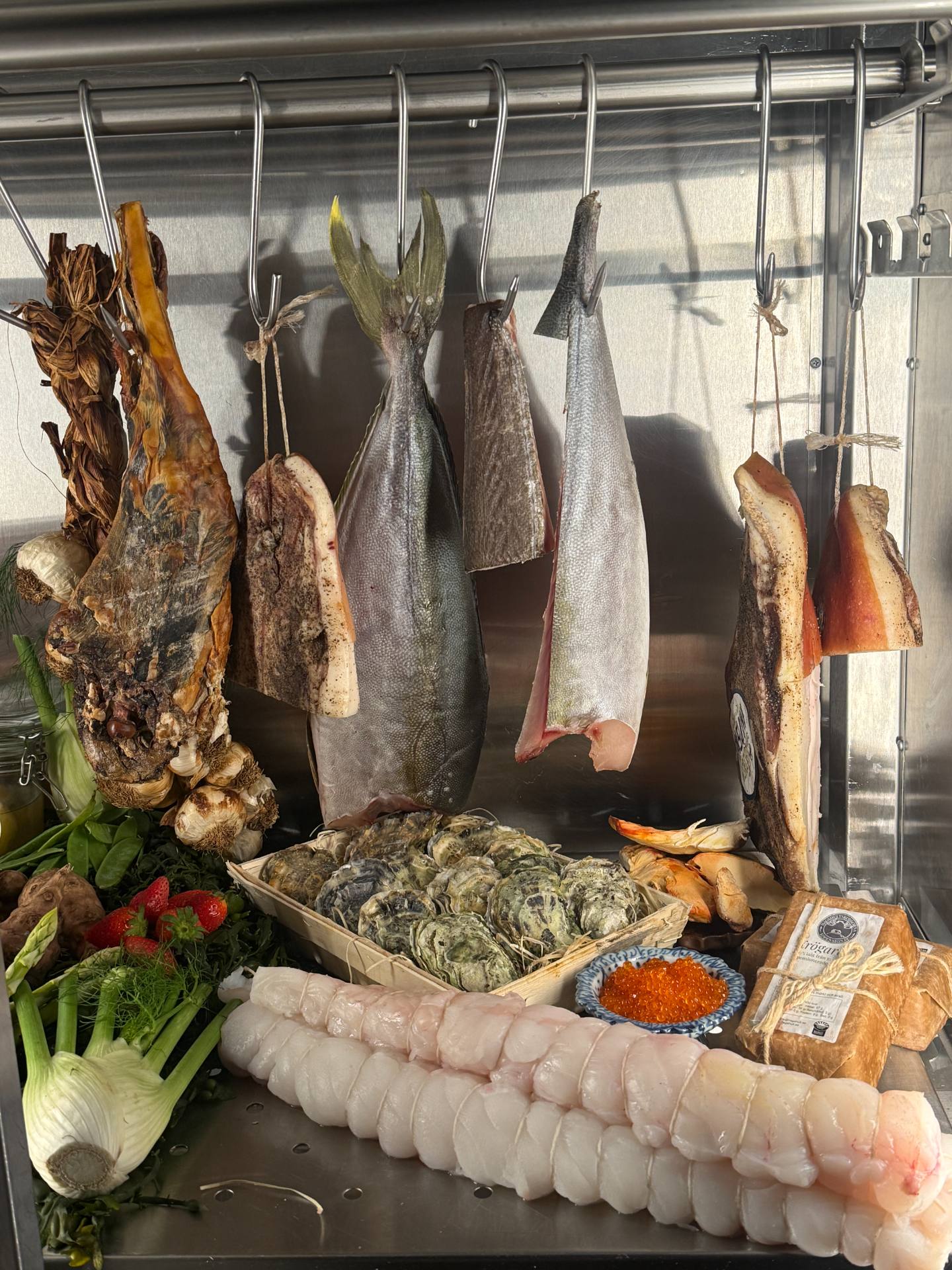 Fish and cuts of meat hanging above oysters, vegetables and roe in Persona’s ingredient fridge.