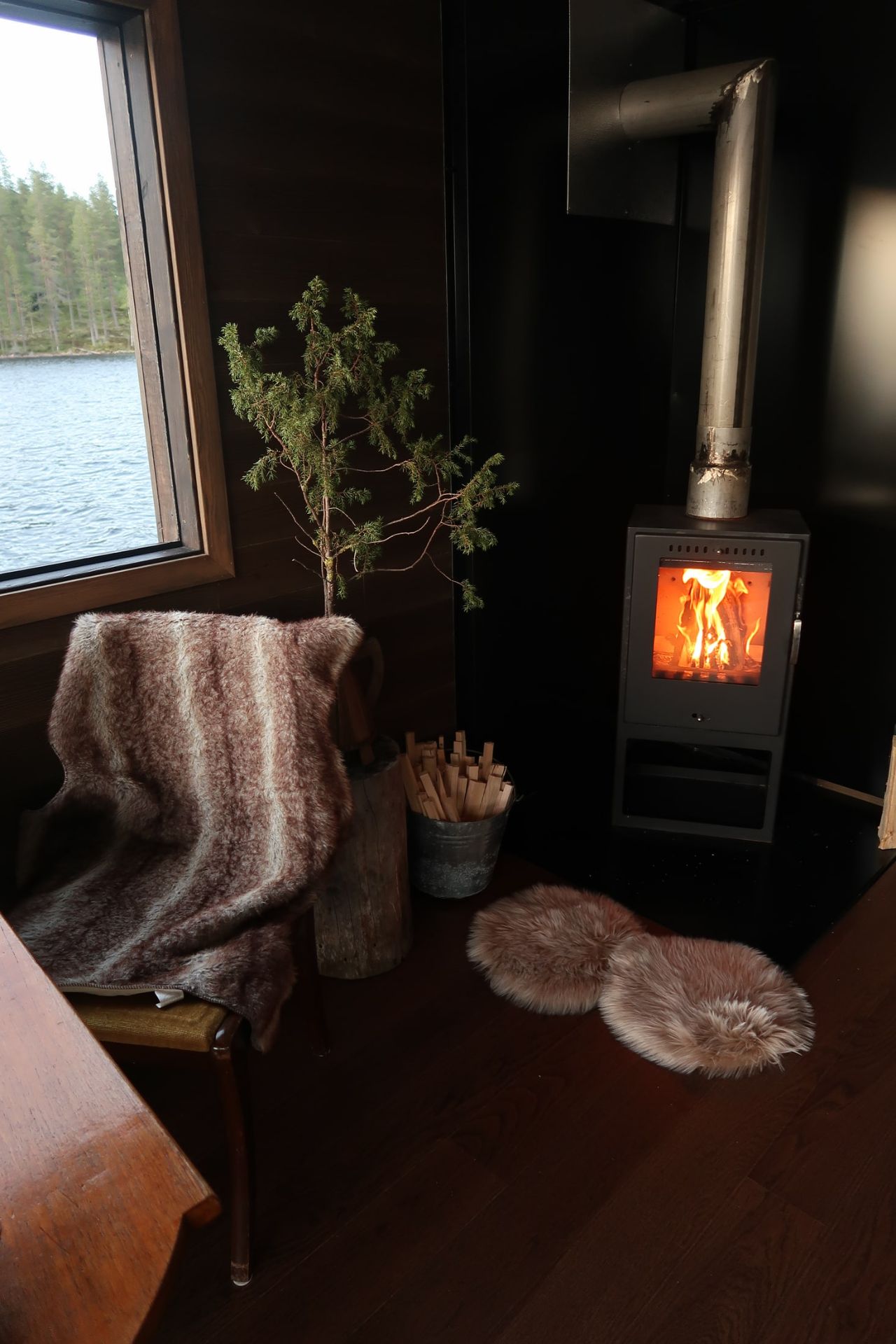 A lit wood-burning stove beside a chair and a window looking across the lake.