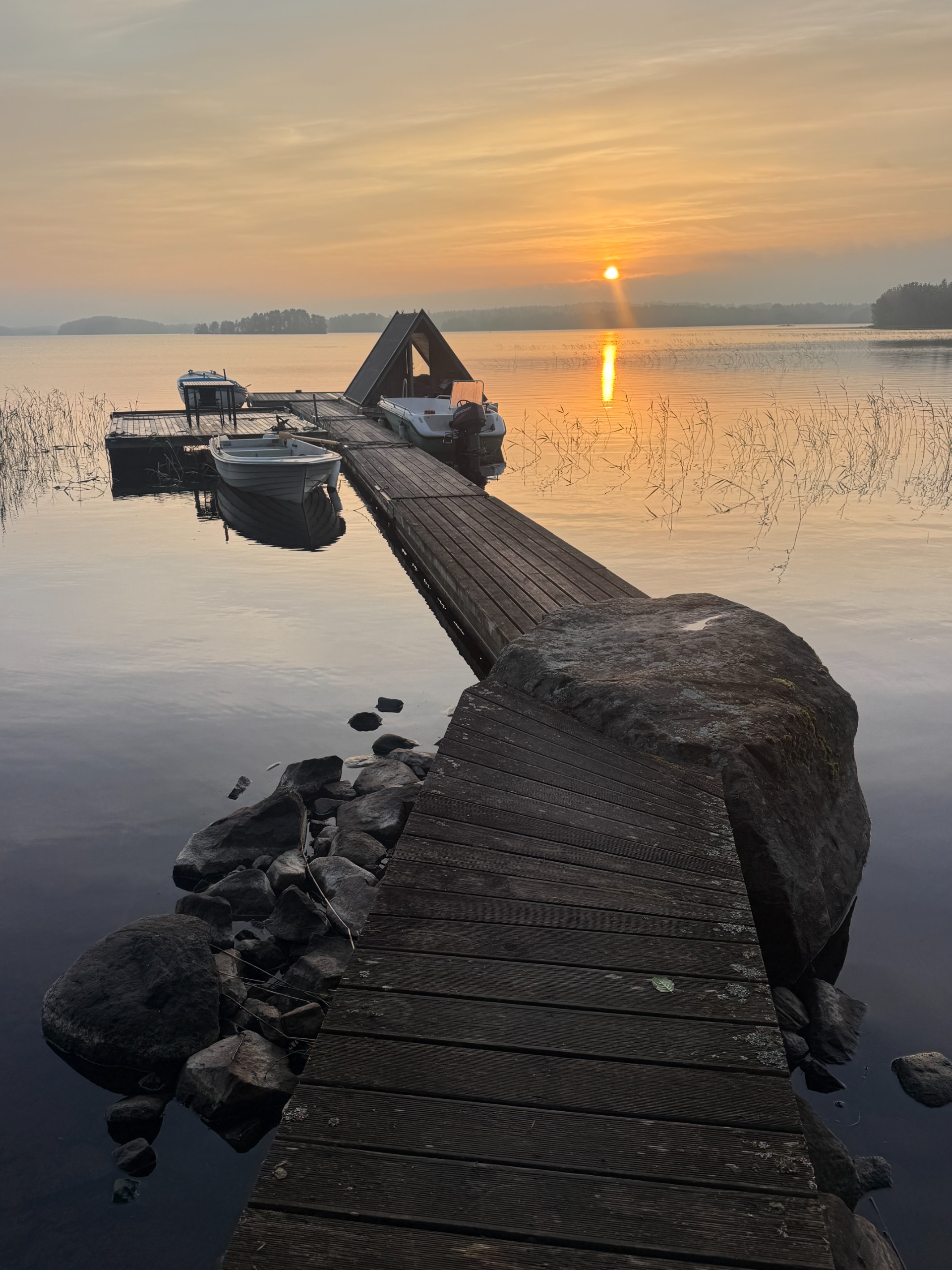 A wooden jetty leading to a tiny floating A-frame cabin and moored boats in golden evening light.