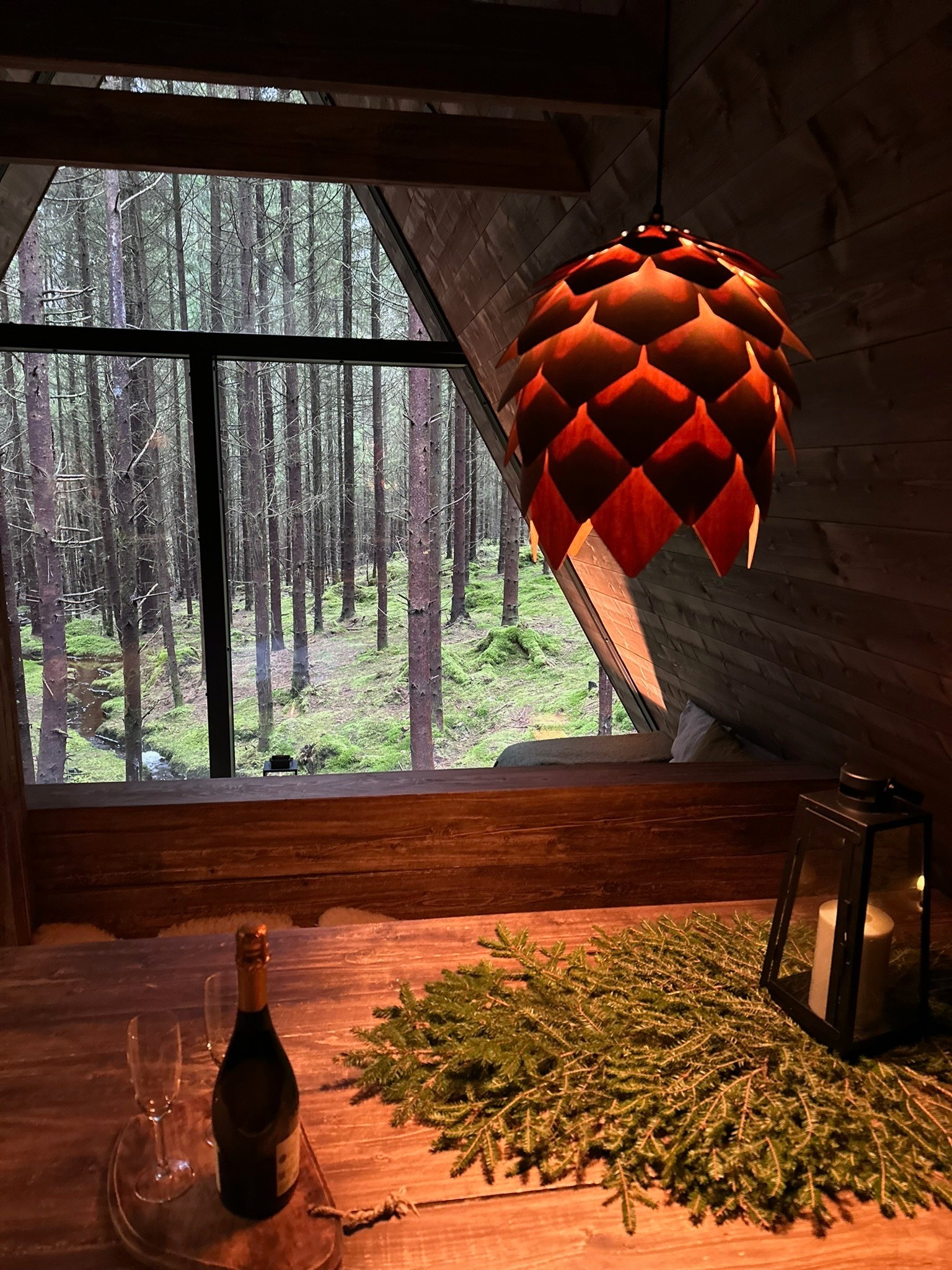 A pine-cone-shaped pendant lamp above a timber table, with forest beyond the window.