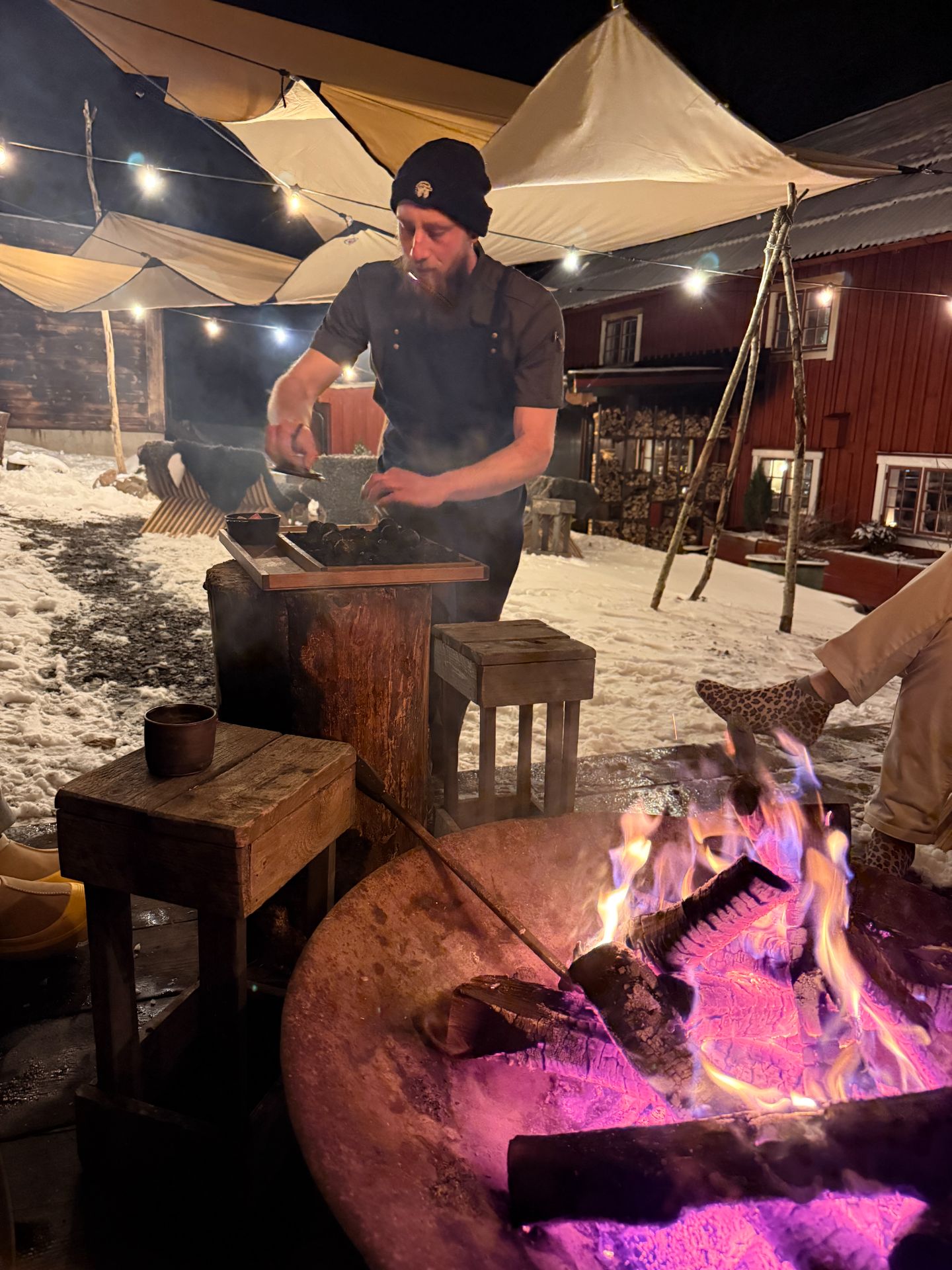 A chef preparing food beside a fire bowl outdoors, with snow, wooden stools and a red timber building behind him.