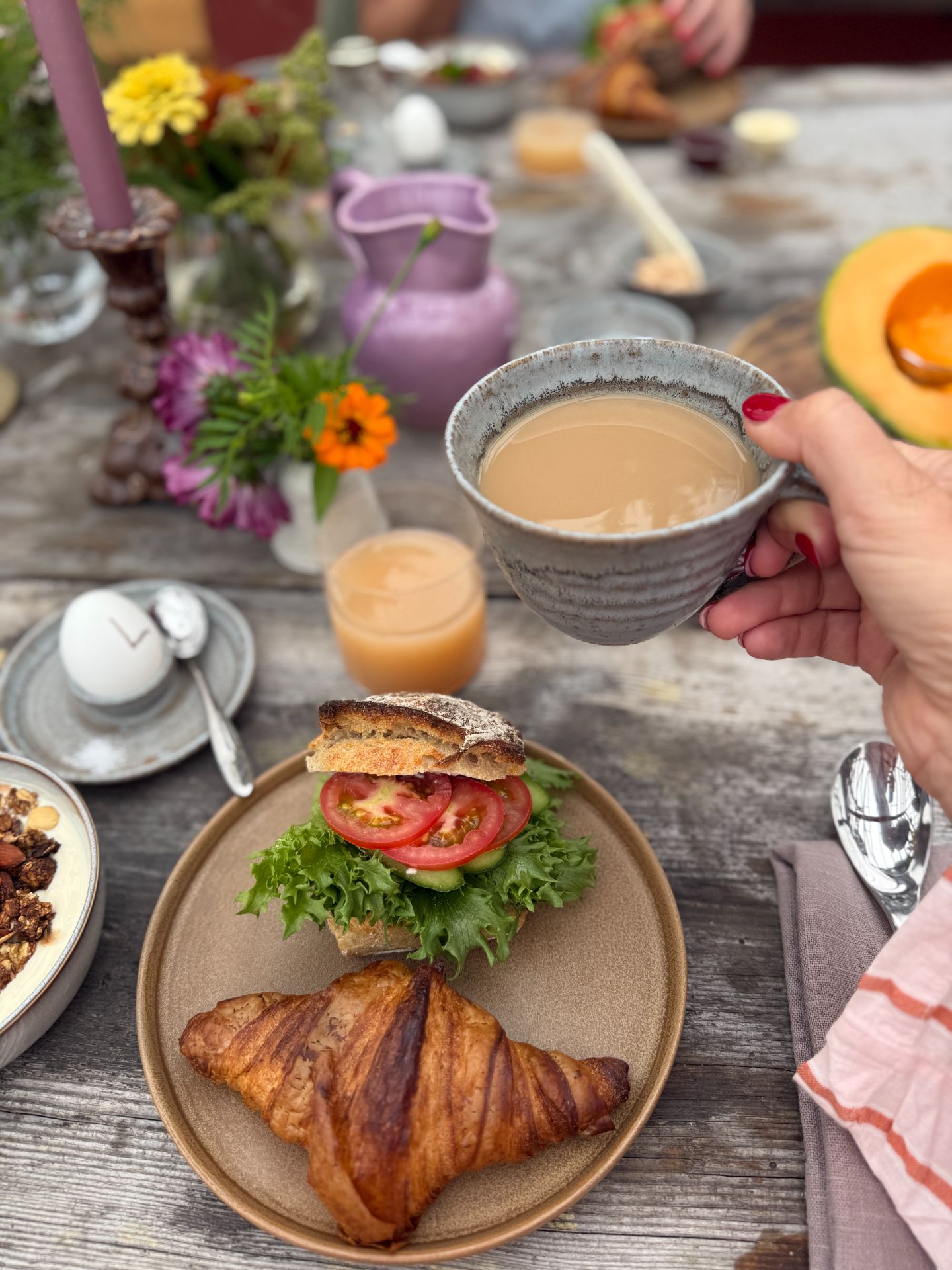 Coffee held above a breakfast table with bread, a croissant and melon.