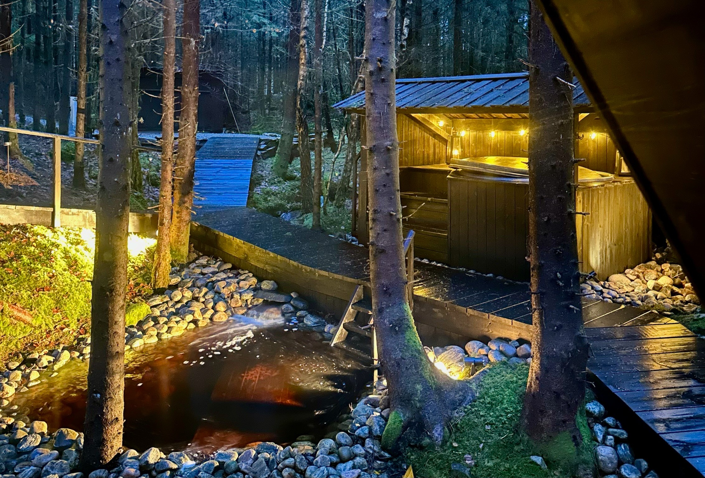 The outdoor hot tub and cold stream beside illuminated wooden walkways at dusk.
