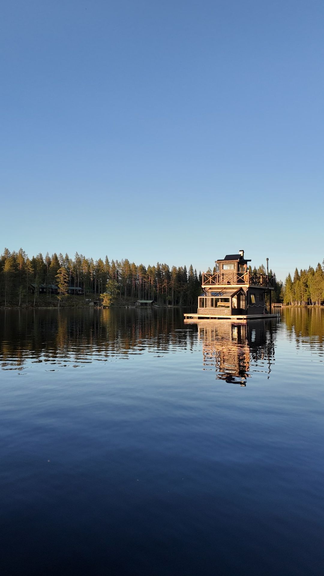 Raven floating on a calm lake, with its wooden cabin and rooftop sauna reflected in the water.