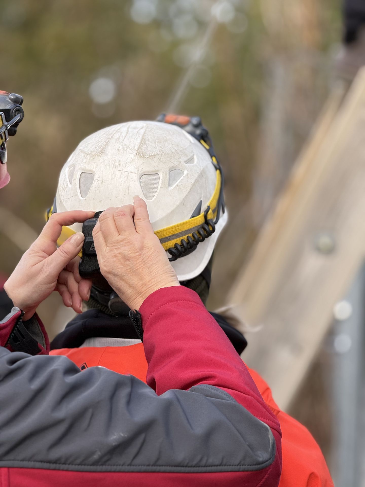 Hands adjust equipment on the back of a white helmet worn by a person in an orange jacket.