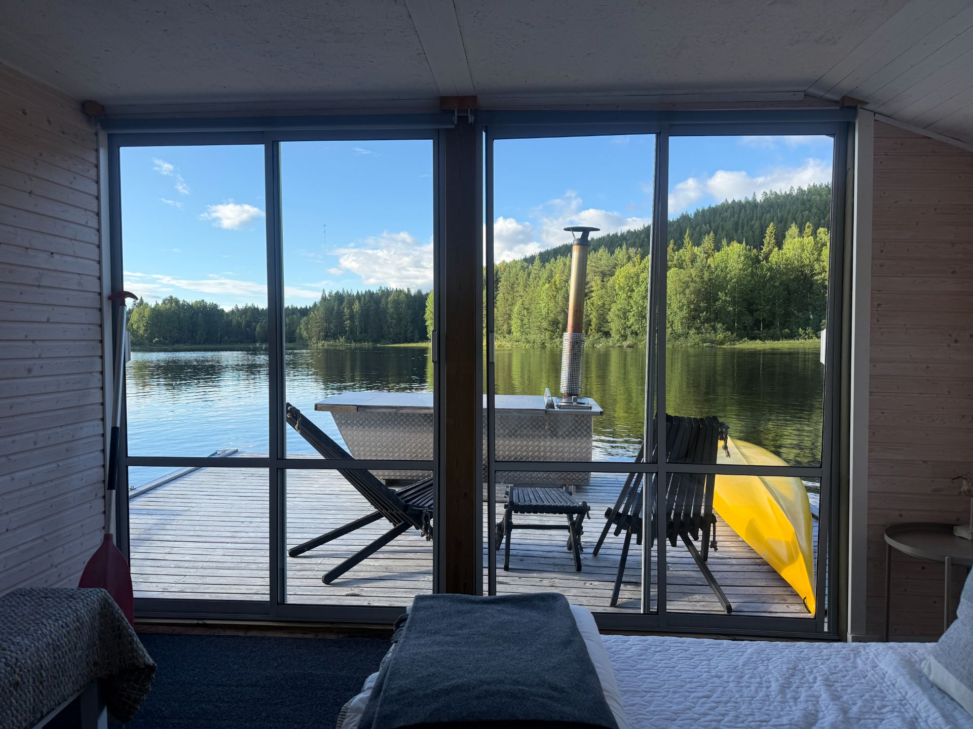 View from the double room through glass doors towards deck chairs, a hot tub and the lake.