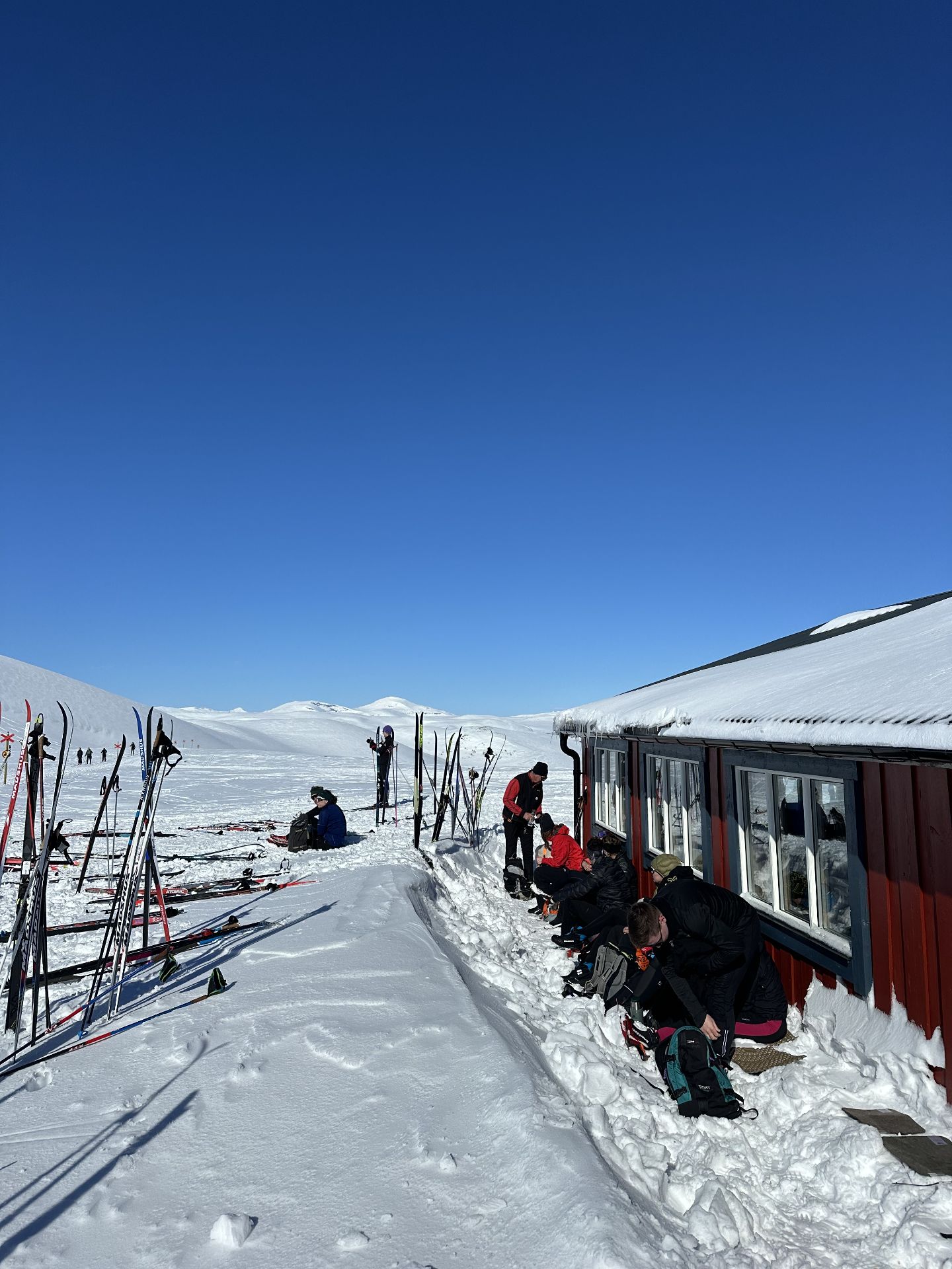 Skiers sit beside a red wooden hut in the snow, with skis and poles standing nearby.