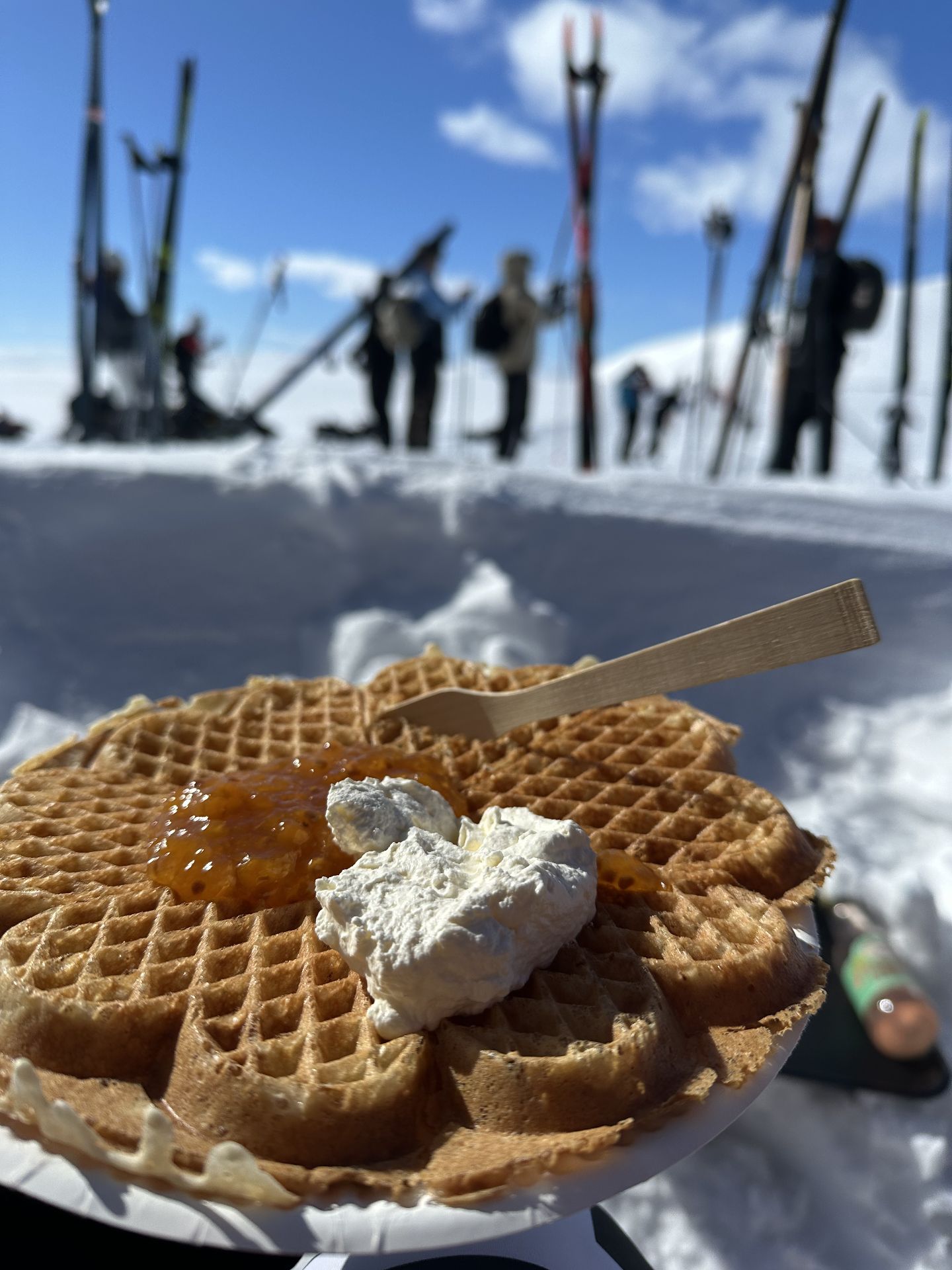 A waffle topped with cream and orange-coloured jam on a paper plate, with skis standing in the snow behind it.