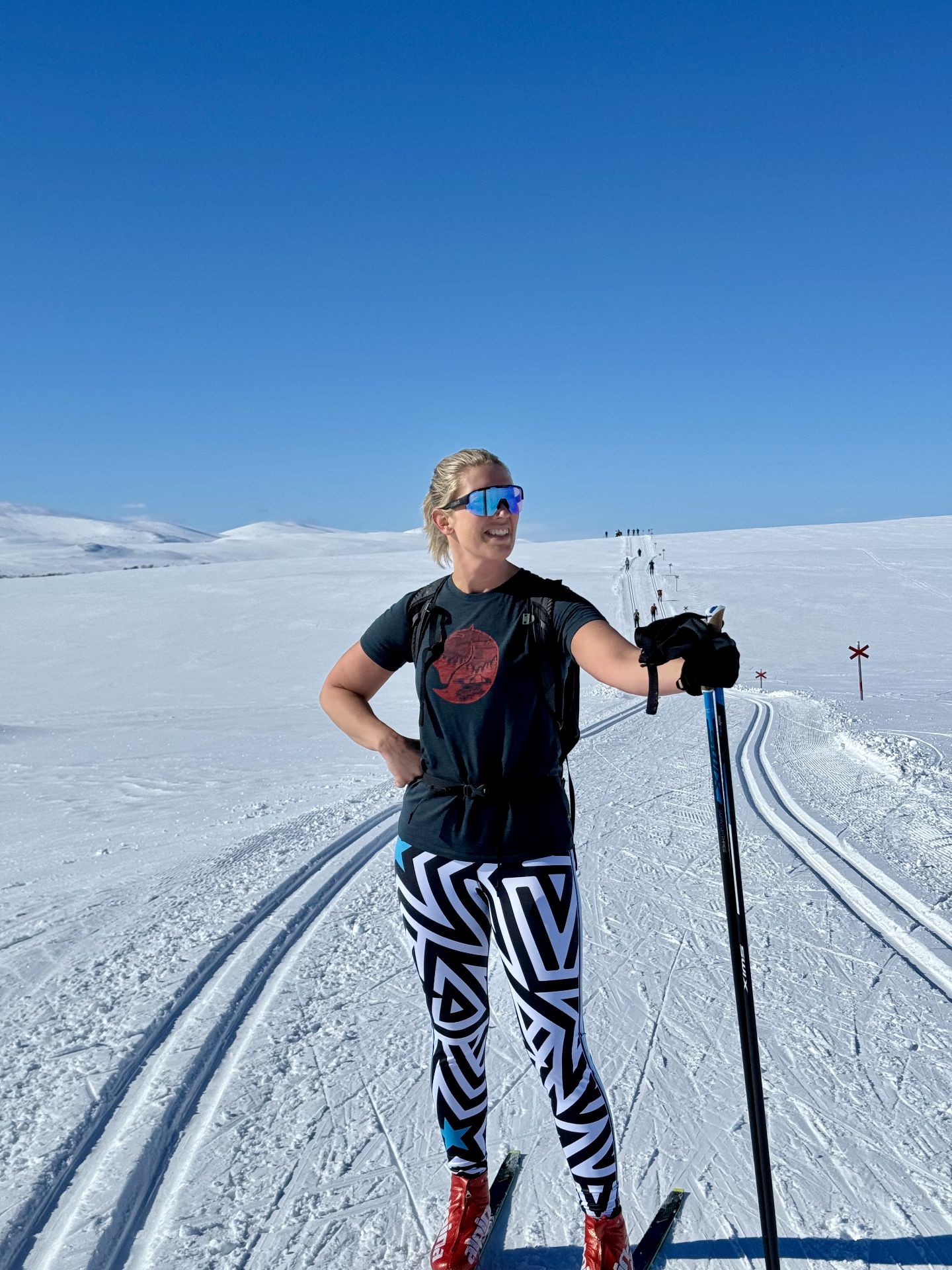 A skier in a black T-shirt, patterned leggings and mirrored sunglasses stands with poles on a snowy trail.