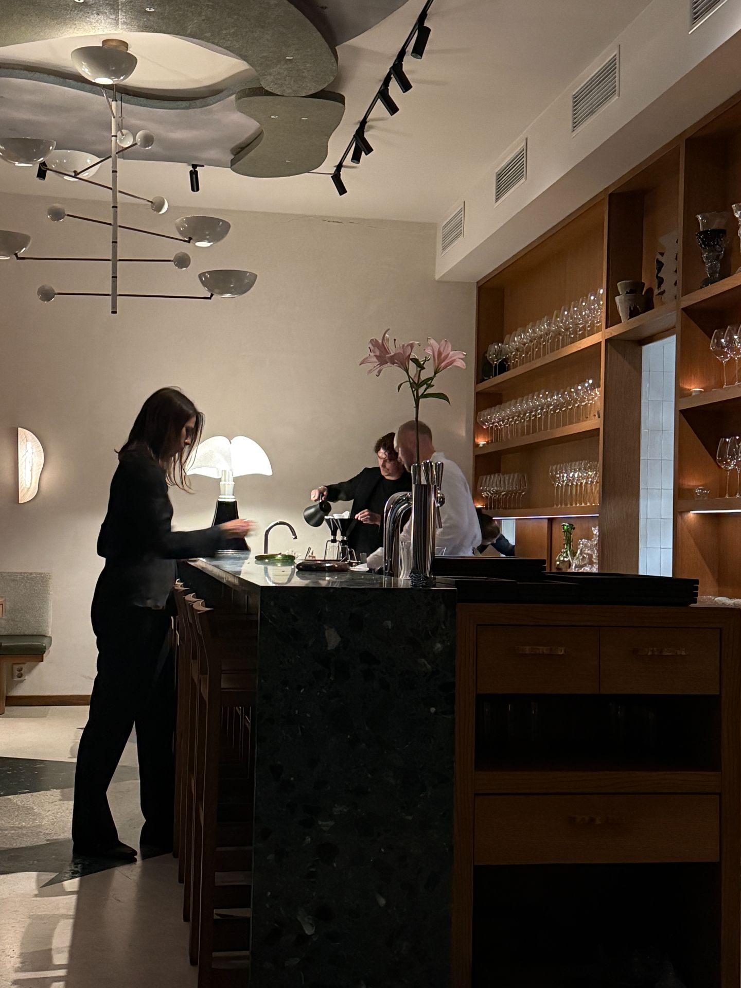 Staff at Persona’s dark stone counter beneath sculptural ceiling lights, with glasses on wooden shelves.