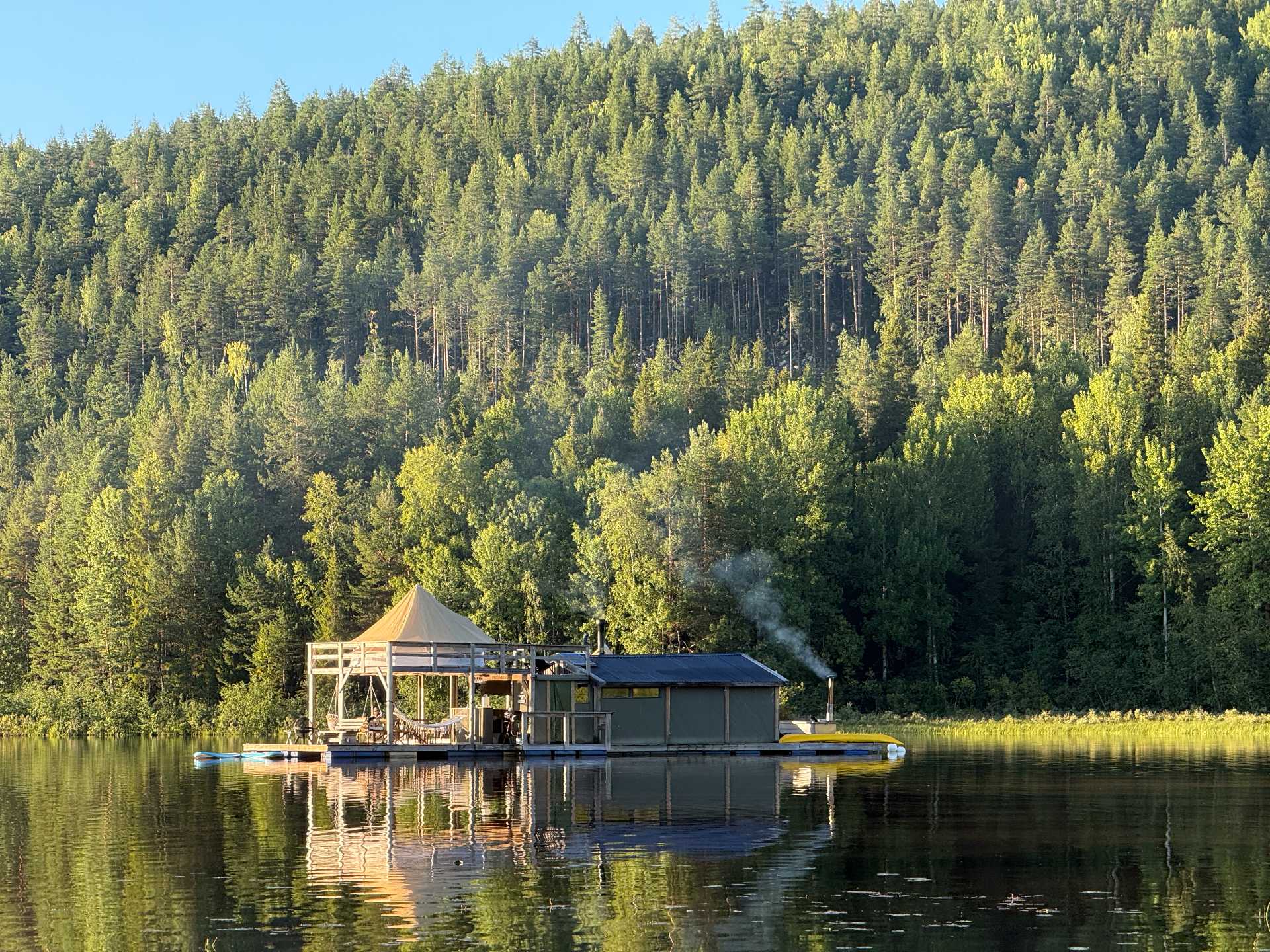 A safari tent and cabin on a floating wooden platform, reflected in a lake beneath a forested hillside.
