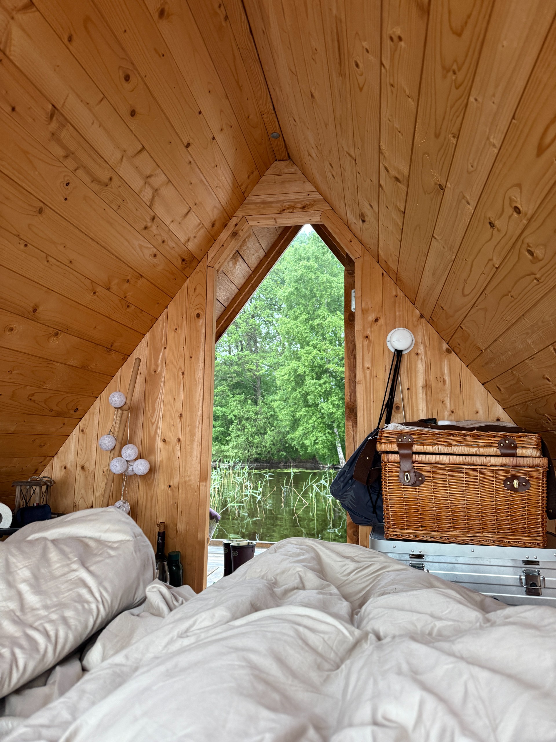 White bedding and a wicker basket inside a timber A-frame cabin, with reeds and trees outside the window.