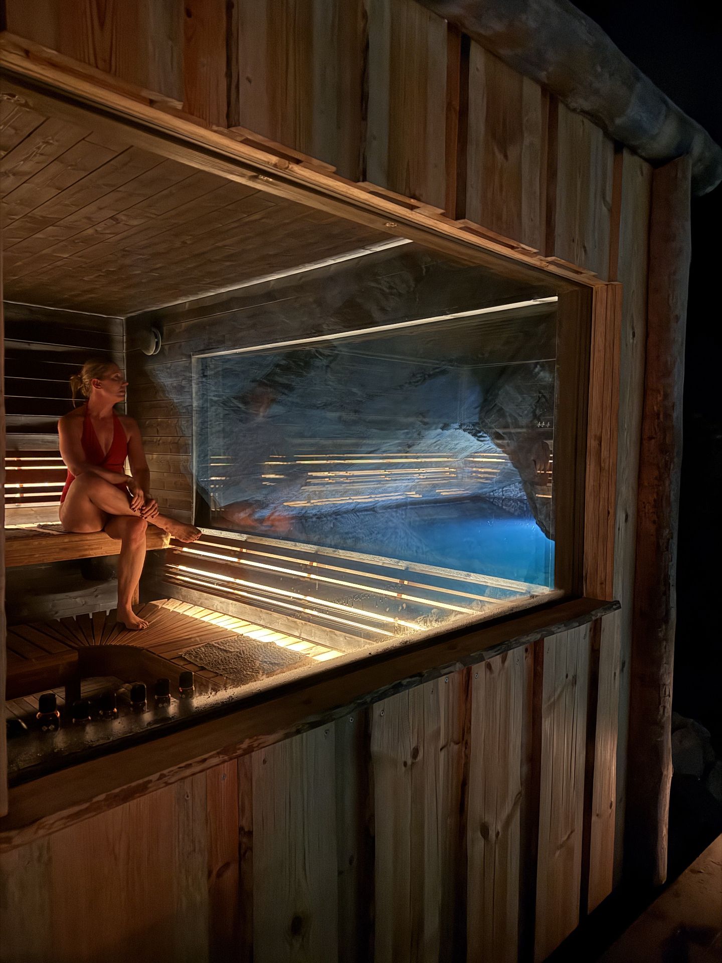 A person in a red swimsuit sits inside a wooden sauna beside a large window overlooking blue mine water and rock.