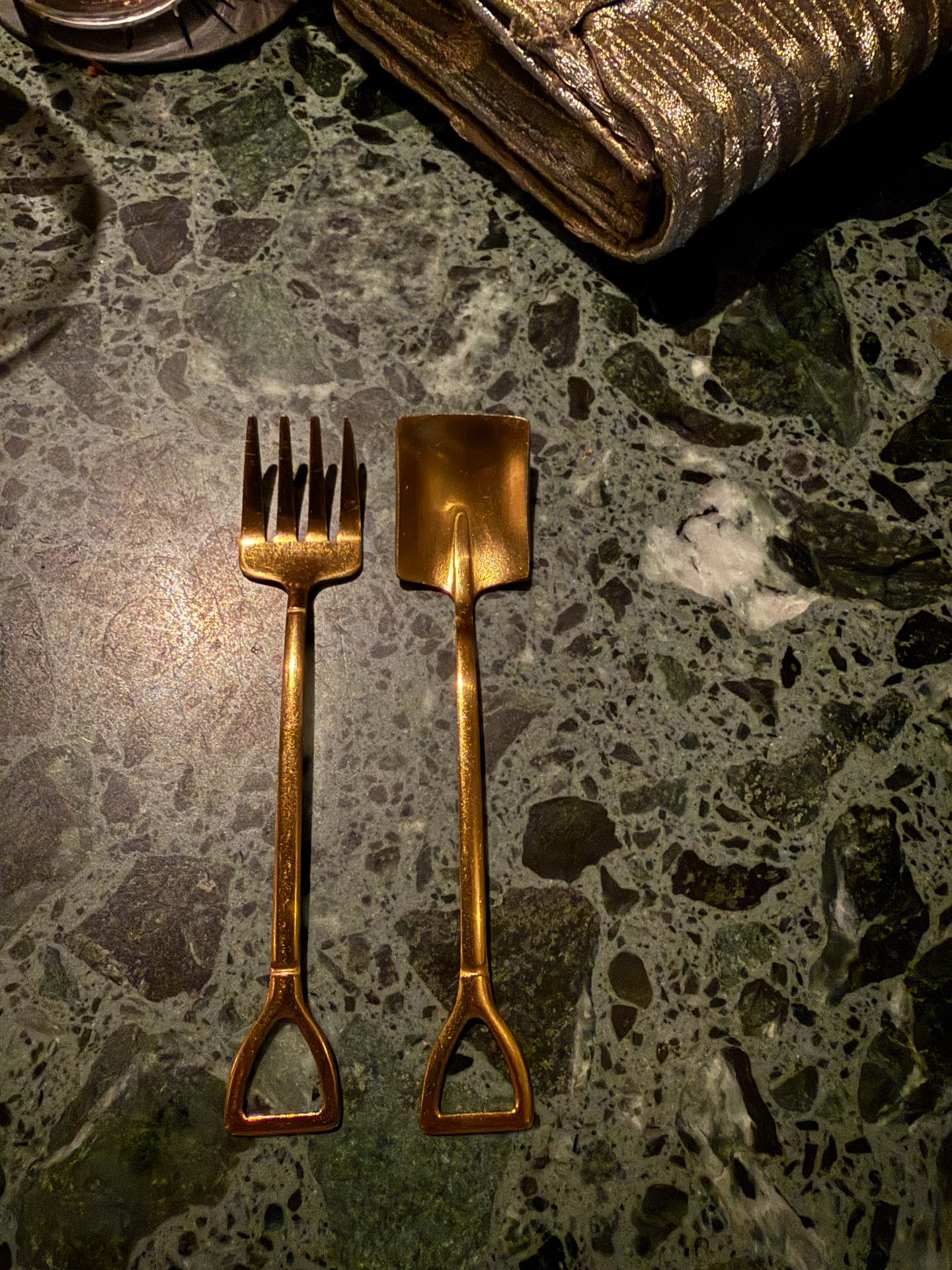 Gold-coloured cutlery shaped like a miniature garden fork and spade on a green stone tabletop.