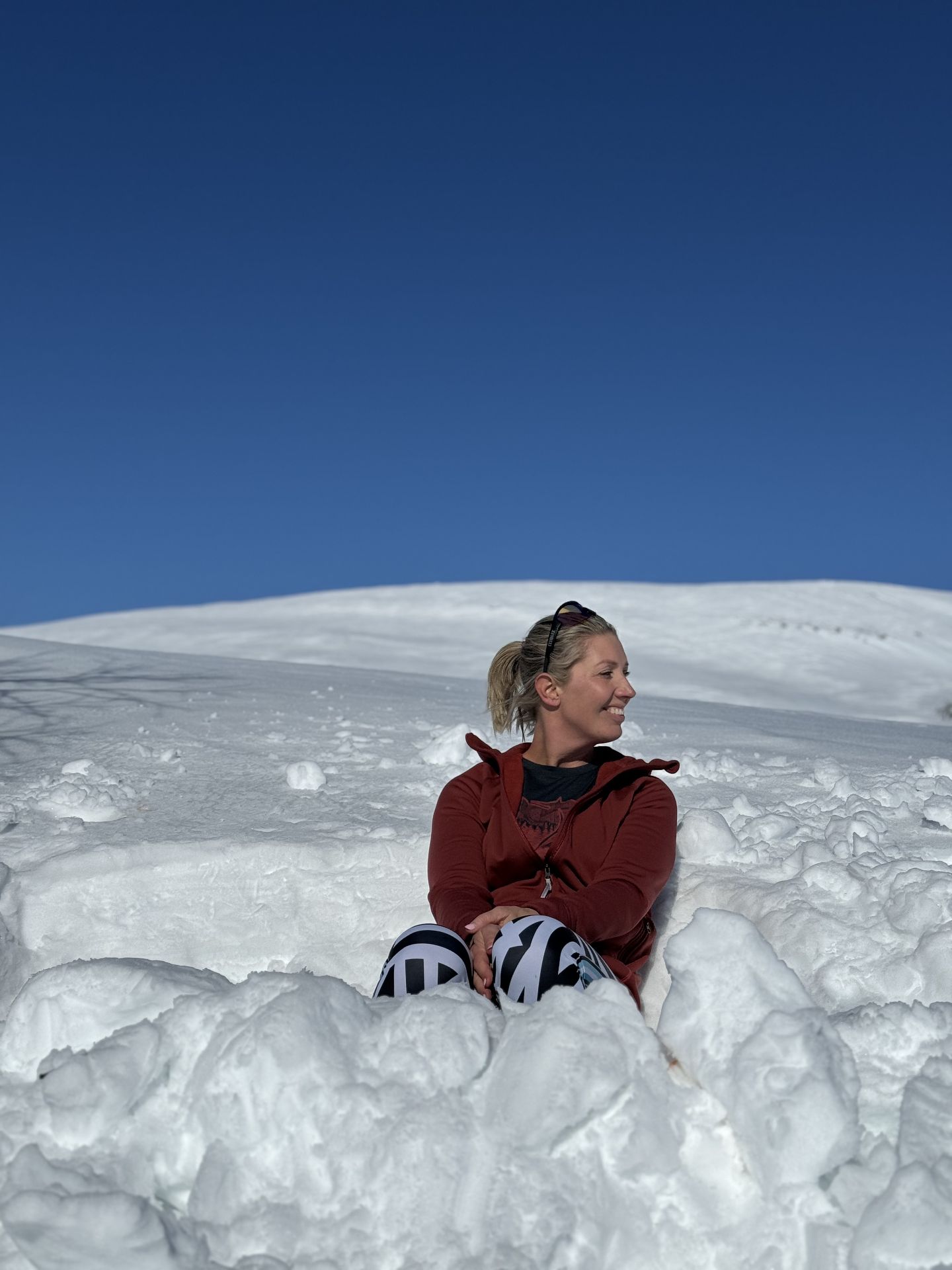 A woman in a red jacket and patterned leggings sits in a hollow in the snow beneath a blue sky.