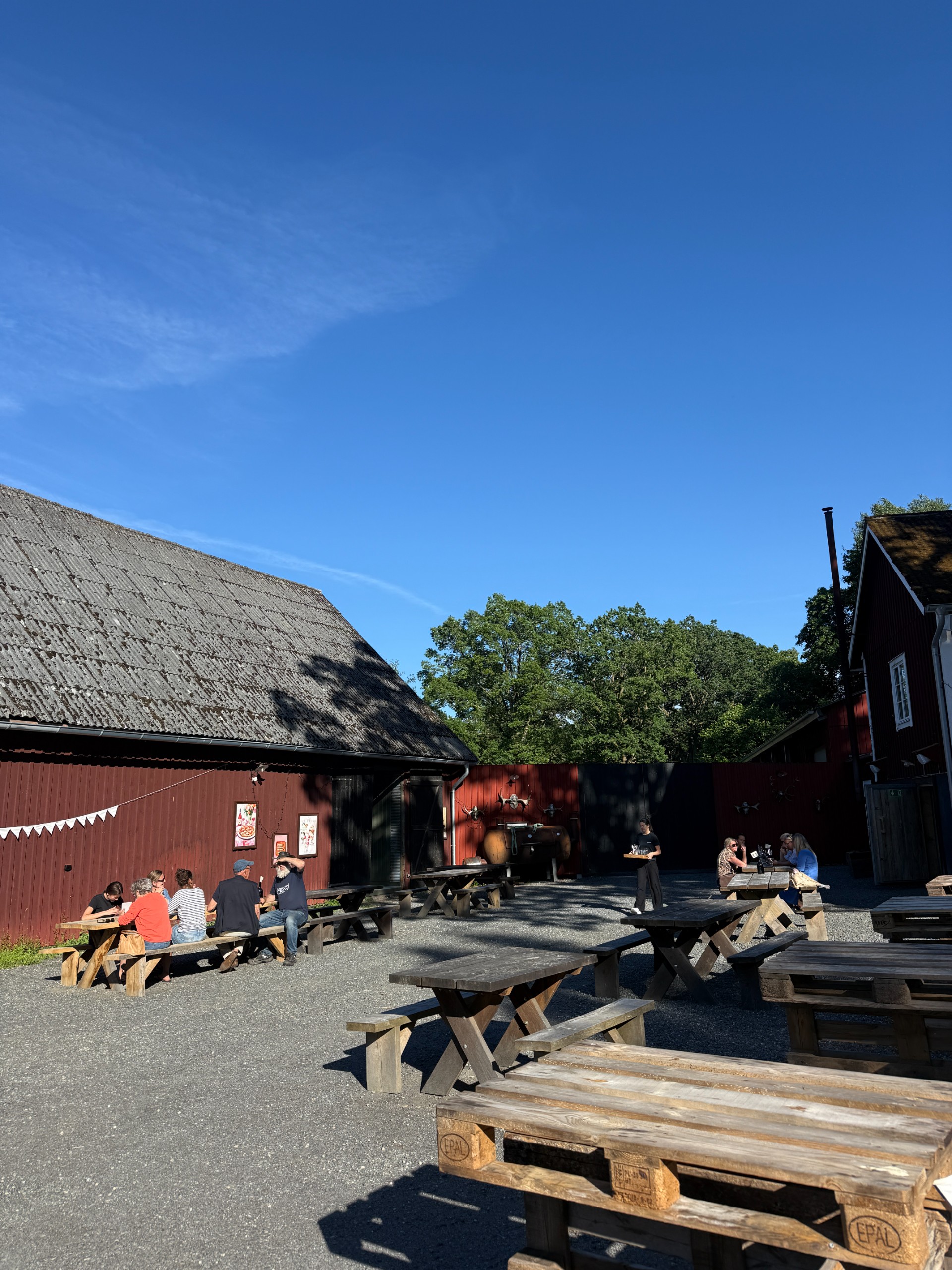 Guests at wooden picnic tables in a courtyard between red farm buildings.