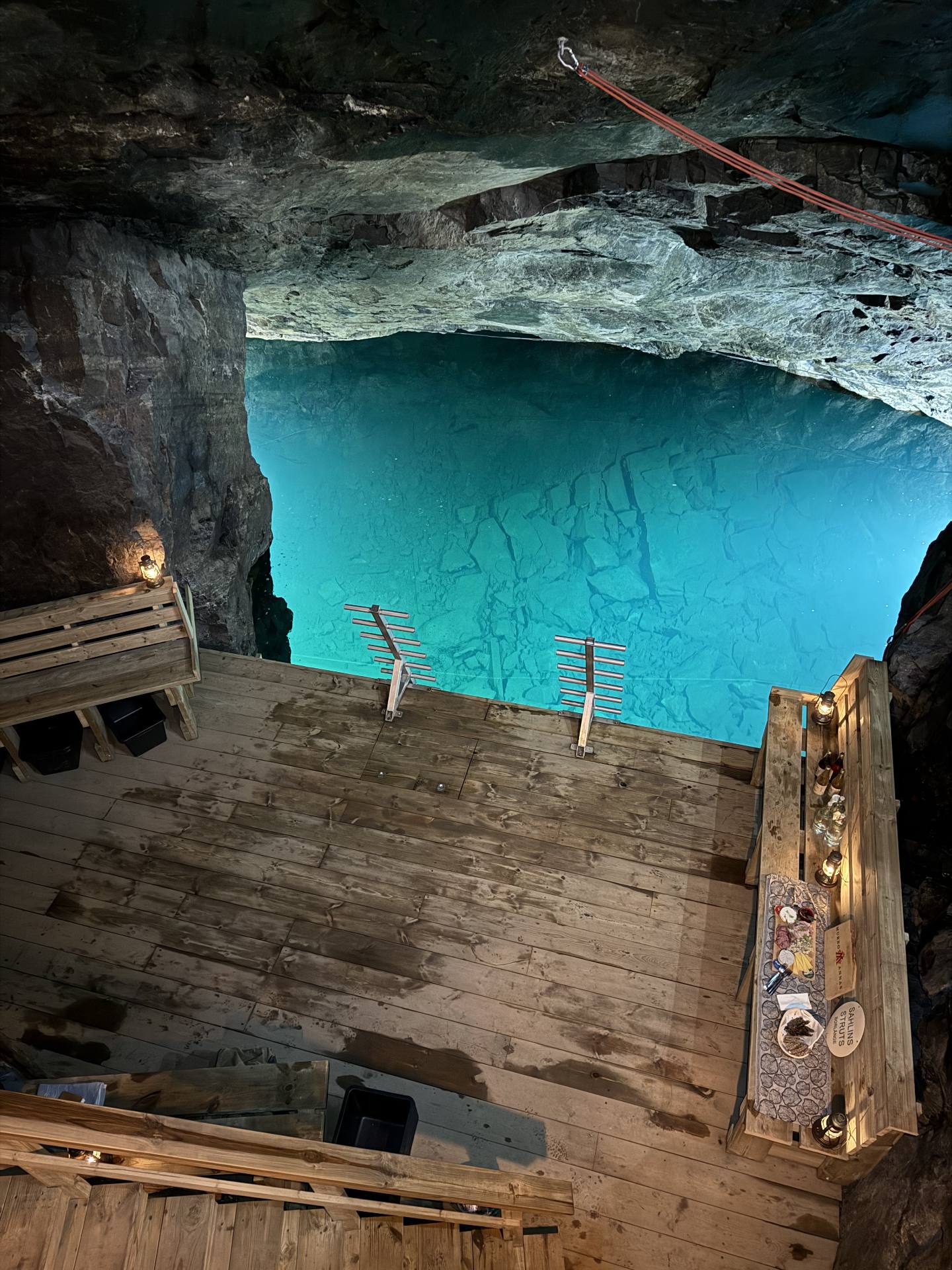 Two ladders descend from a wooden platform into clear blue mine water beneath a rock ceiling.