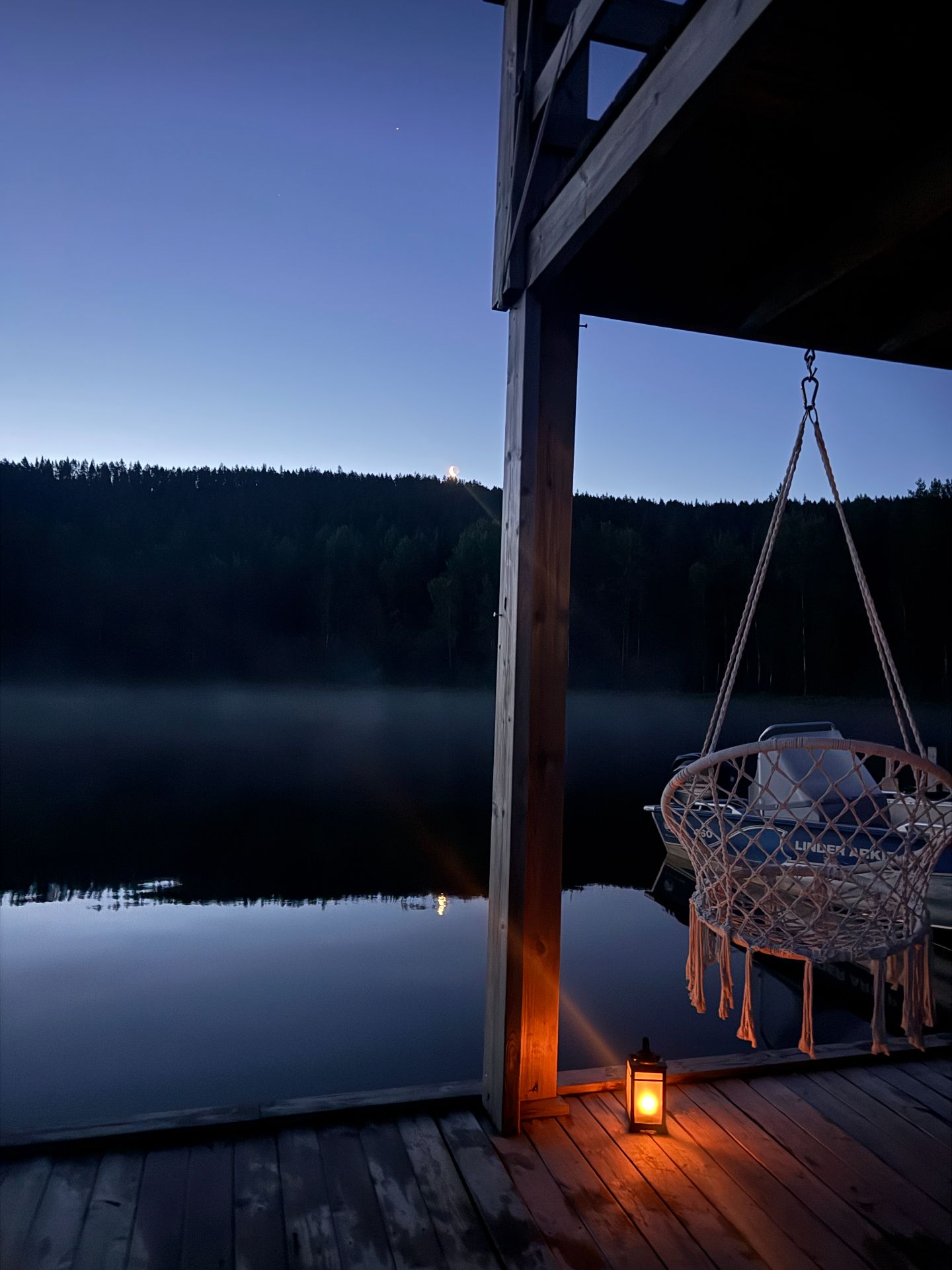 A hanging chair and glowing lantern on the deck beside a misty lake at night.